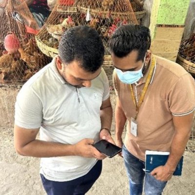 Trainer holding a cellphone, and explaining an app to a trainee, in a live market, with poultry in the background.