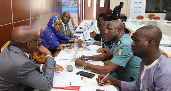 Seven people sitting around a long table with numerous documents during a working session.