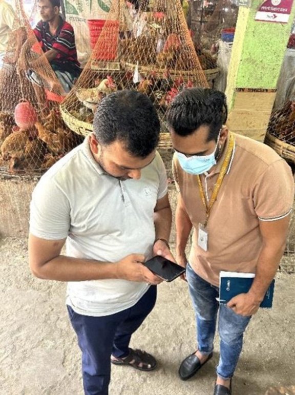 Trainer holding a cellphone, and explaining an app to a trainee, in a live market, with poultry in the background.